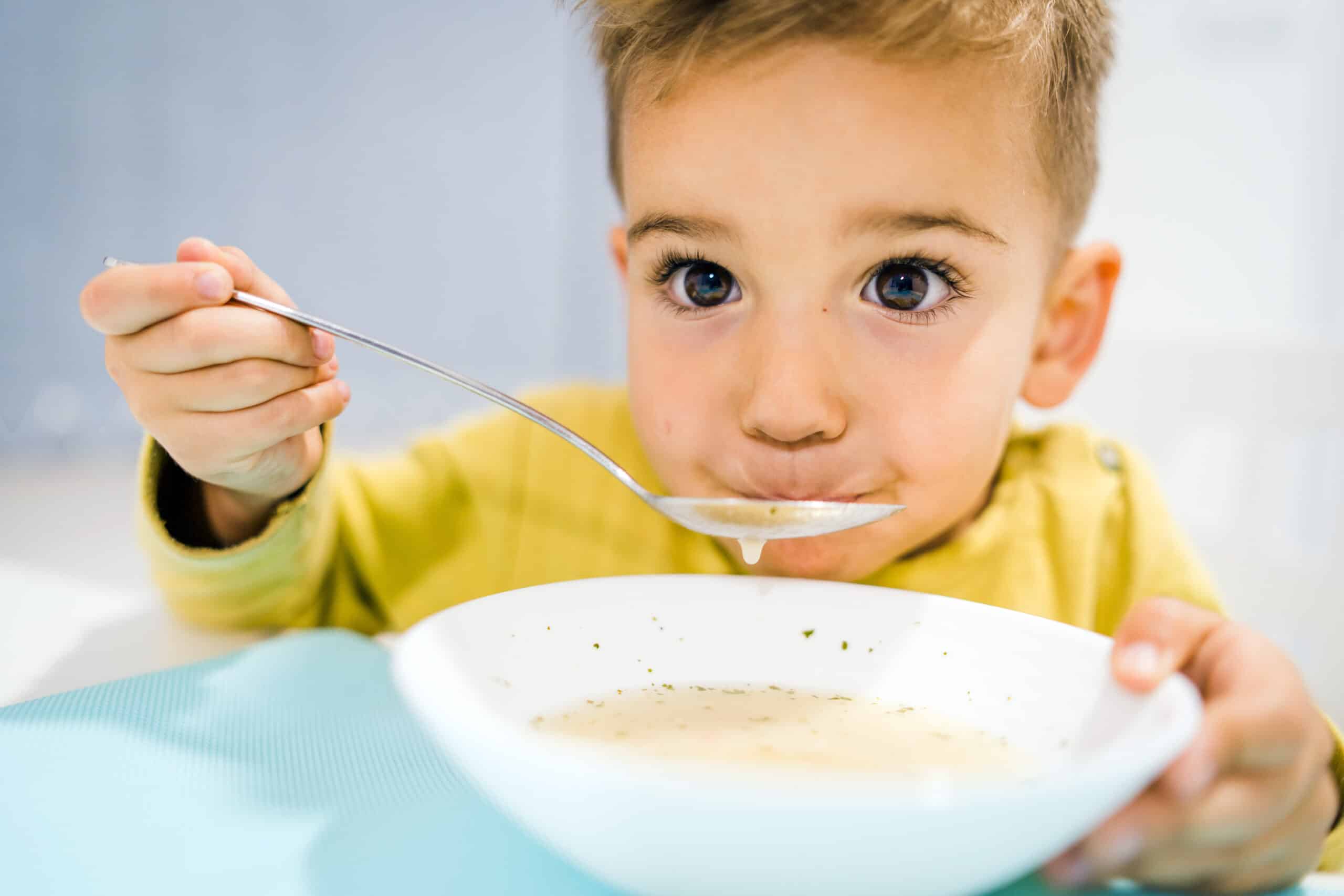 Portrait of small boy child eating soup meal or breakfast having lunch by the table at home with spoon white